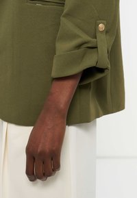 Bras à la peau foncée avec une manche olive retroussée et maintenue par un bouton doré, sur un fond de pantalon crème et mur blanc.