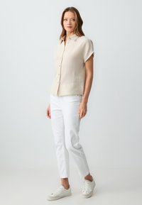 Donna che indossa una camicia beige con maniche corte e bottoni, pantaloni bianchi fino alla caviglia e sneakers bianche, in piedi contro uno sfondo chiaro e semplice.