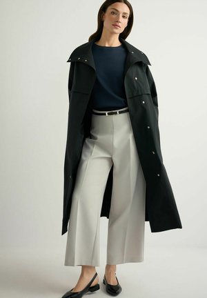 Kobieta ubrana w czarny długi płaszcz, granatową bluzkę, jasnoszare szerokie spodnie z paskiem oraz czarne buty na obcasie, stojąca na jednolitym tle.