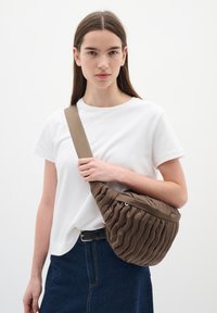 Borsa a tracolla marrone trapuntata con forma curva e superficie texturizzata. Cinghia regolabile e dettagli hardware discreti. Indossata con una maglietta bianca.