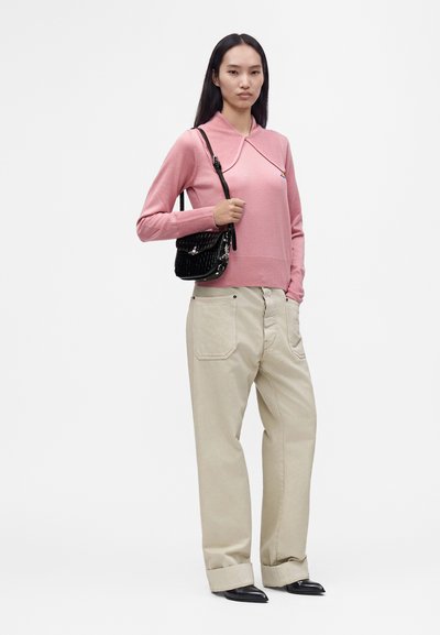 Femme portant un haut rose à manches longues, un pantalon beige à jambes larges, des chaussures noires pointues, tenant un sac à bandoulière noir texturé.