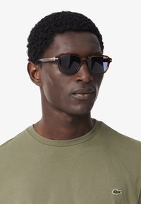 Occhiali da sole con forma rettangolare, montatura a tartaruga, lenti scure e dettagli metallici sulle aste; indossati con una maglietta verde oliva.