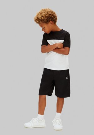 Ragazzo con capelli ricci che indossa una maglietta bianca e nera, pantaloncini neri, calzini bianchi e scarpe da ginnastica bianche, in piedi con le braccia incrociate, guarda verso il basso.