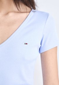 Világoskék csíkozott póló, mély V-kivágással és rövid ujjal. Kis hímzett logó a mellkas közelében. Puha pamut anyag.
