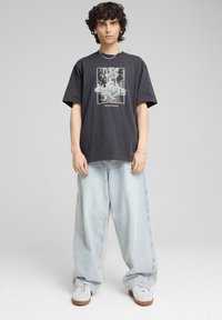 Camiseta gráfica gris de corte relajado, con una impresión blanca de una figura corriendo. Combinada con vaqueros anchos de color azul claro y zapatillas blancas.