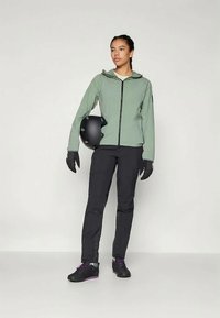 Hele roheline jakk kapuutsiga, musta tõmblukuga, mustad kindad, mustad püksid ja mustad jalatsid lilla aktsendiga, hoides käes musta kiivrit.