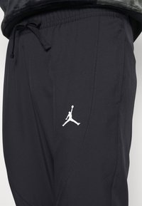 Schwarze Sporthosen aus glattem Material, mit einem verstellbaren Bund und einem weißen Jumpman-Logo auf der linken Hüfte. Rückentasche sichtbar.