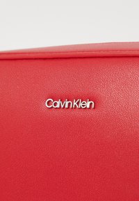 Rote Handtasche aus Kunstleder mit glatter Textur, mit einem silbernen "Calvin Klein"-Logo, minimalistisches Design und subtilen Steppdetails.