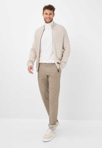 Beige geribbelde ritscardigan over een witte turtleneck, gecombineerd met beige textuurbroek en lichtgekleurde sneakers, poserend tegen een witte achtergrond.