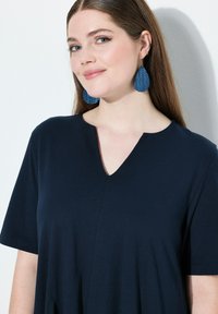 Top azul marino de manga corta con escote en V y corte relajado. Presenta una textura sutil y un pequeño detalle de costura en el frente. Pendientes en forma de lágrima en un azul contrastante.
