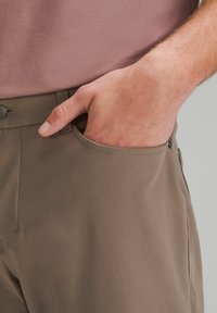 Ruskeat housut, joissa on sileä, teksturoitu kangas. Etutasku, johon käsi on asetettu. Napillinen kiinnitys vyötäröllä.