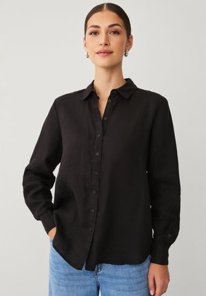 Donna con capelli scuri che indossa orecchini a cerchio argento, una camicia nera abbottonata e jeans azzurri, in piedi con una mano in tasca.