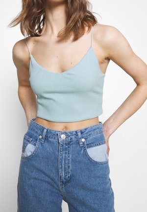 Frau trägt ein hellblaues, geripptes Crop-Top mit dünnen Trägern und hochtaillierte blaue Jeans mit aufgesetzten Taschen, steht mit hinter dem Rücken verschränkten Händen.