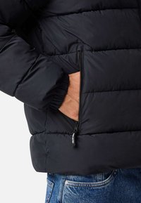 Czarna kurtka puchowa z pikowanymi segmentami, elastycznymi mankietami i kieszenią na zamek. Nosi się ją na niebieskich dżinsach z widocznym pasem.