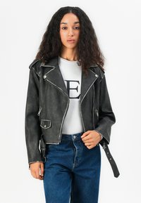 Black faux leather biker jacket with silver hardware, zippers, and a pointed collar. Worn over a white shirt with a large black letter.