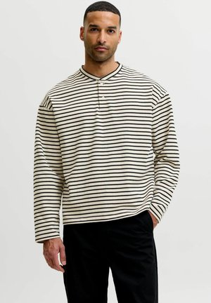 Mann mit kurzen Haaren und gestutztem Bart, der ein cremefarbenes, langärmeliges Hemd mit schwarzen horizontalen Streifen und schwarze Hose trägt, steht mit der Hand in der Tasche.