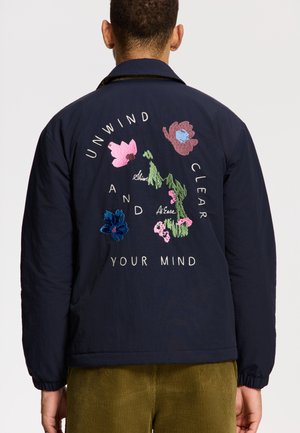Marineblauwe geborduurde jacket met bloemontwerpen en de tekst "ONTSPAN EN CLEAR UW GEEST." Lichtgewicht stof met elastische manchetten.