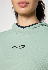 Mintgrønn hettegenser med en teksturert overflate og et svart uendelighetssymbol på brystet. Marineblå kant rundt halsen fullfører designet.