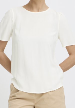 Woman wearing a plain white short-sleeve blouse tucked into beige pants, standing with hands behind her back.