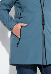 Chaqueta impermeable azul con una superficie texturizada, que cuenta con una cremallera frontal, bolsillos laterales y un cinturón en la cintura para un ajuste ajustable.