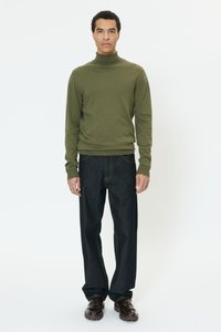 Olivno zelena rebrasta pulover z visokim ovratnikom in oprijetimi rokavi, kombiniran s temno modrimi kavbojkami z ravnimi hlačnicami in temno rjavimi čevlji z vezalkami.