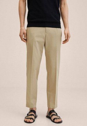 Modèle masculin portant un pantalon beige sur mesure et des sandales noires, debout devant un fond clair uni.