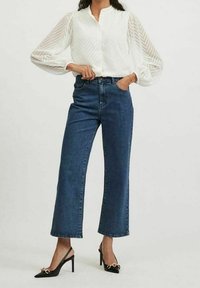 Valkoinen liivihihainen paita, jossa on teksturoitu, savupiippukuvioinen hihapari, yhdistettynä korkea vyötäröisiin, suoriin sinisiin farkkuihin ja mustiin avokkaisiin, joissa on kultaiset yksityiskohdat.