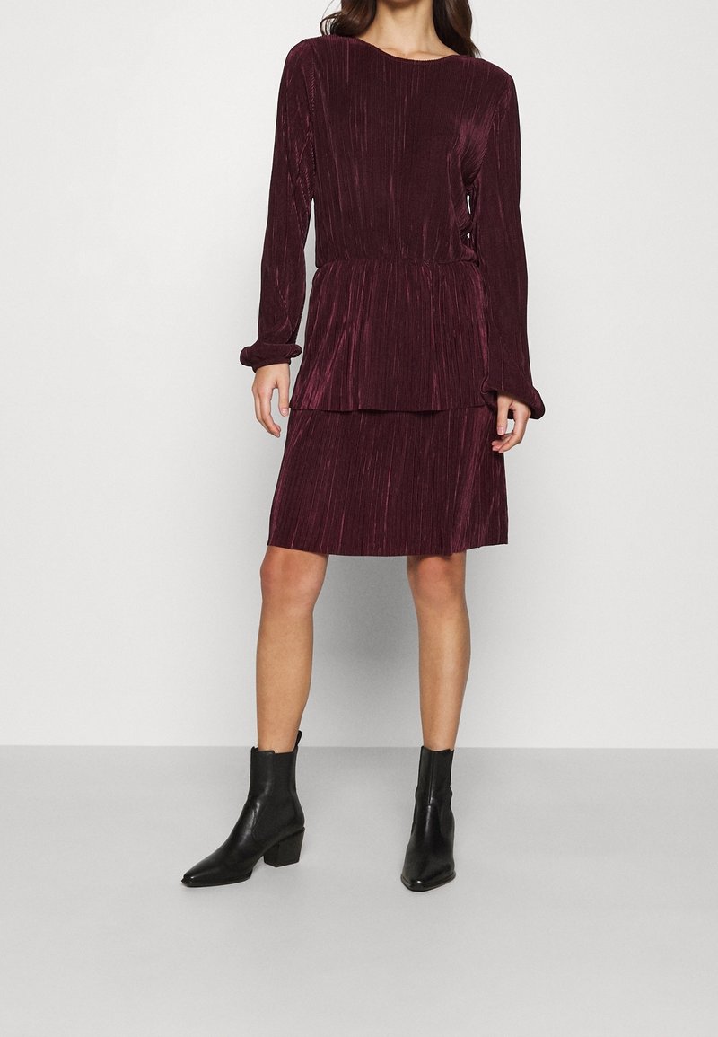 Burgunderfarbenes, plissiertes Kleid mit langen Ärmeln, tailliertem Bund und geschichtetem Saum. Kombiniert mit schwarzen Lederstiefeletten.