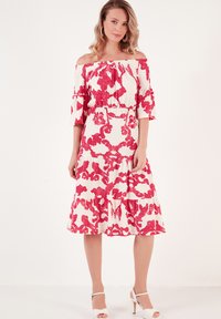 Off-the-shoulder dress in a white fabric with bold red floral patterns, flared sleeves, and a tiered hemline. Features a fitted waist.