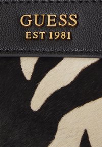 Material de estampado animal en negro y beige con una textura suave, que presenta un logo en relieve dorado que dice "GUESS EST 1981" en una banda de cuero.