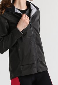 Mujer vestida con una chaqueta negra con cremallera sobre una camiseta gris oscuro y pantalones cortos de ciclismo con paneles rojos, posando frente a un fondo sencillo.
