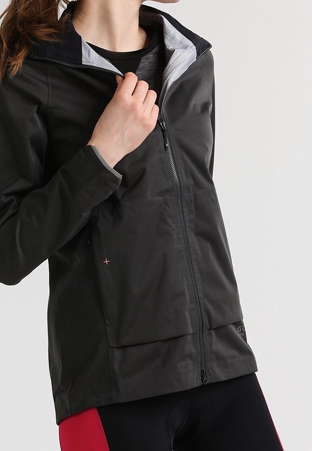 Mujer vestida con una chaqueta negra con cremallera sobre una camiseta gris oscuro y pantalones cortos de ciclismo con paneles rojos, posando frente a un fondo sencillo.