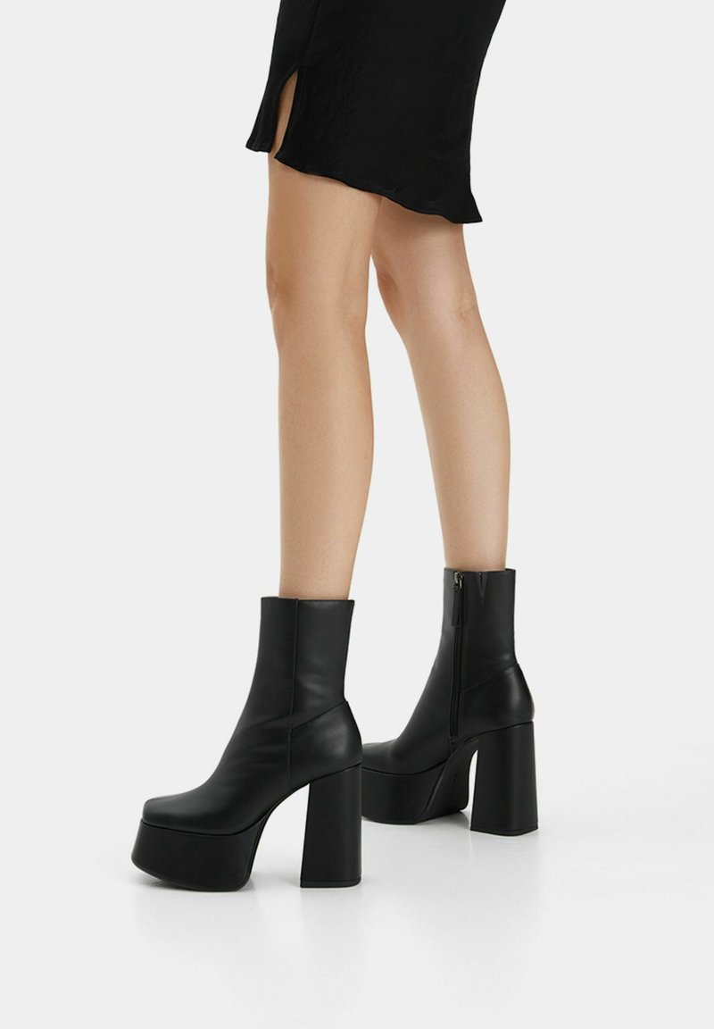 Piernas en botas tobilleras negras de plataforma con tacones gruesos y cremallera lateral, vistiendo una falda negra con una abertura lateral, de pie sobre un suelo blanco.