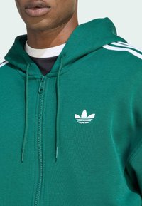 Felpa verde con zip in misto cotone, con accenti bianchi a tre strisce sulle spalle e un piccolo logo bianco sul petto.
