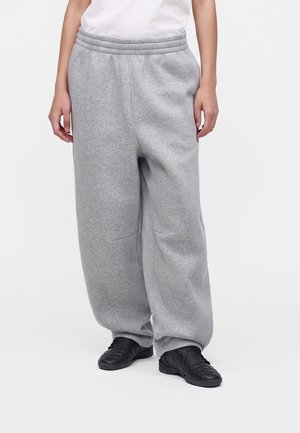 Person iført løse lysegrå joggingbukser og sorte atletiksko står mod en ensartet hvid baggrund.