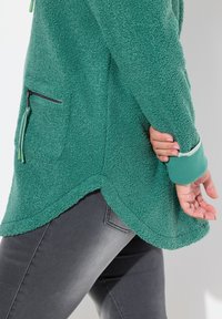 Veste en polaire verte avec une surface texturée, un ourlet arrondi et une poche zippée. Détail contrasté sur les poignets. Associée à un jean gris foncé.