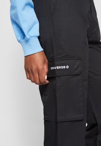 Main reposant sur la poche d'un pantalon cargo noir avec le logo blanc Converse, portant un haut à manches longues bleu clair.