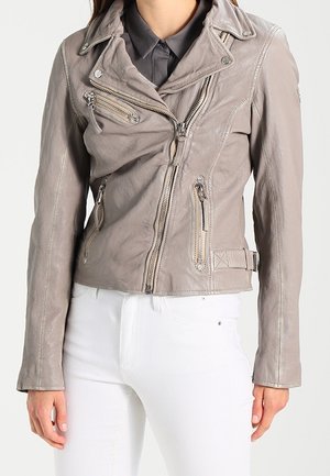 Femme portant une veste biker en cuir gris clair avec plusieurs poches zippées et un pantalon blanc sur un fond uni.