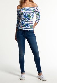 Schulterfreies Oberteil mit blauen Blumenmustern auf weißen Streifen, kombiniert mit enger blauer Jeans und weißen Sneakern.