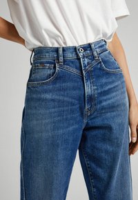 Person som bär blå högmidjade jeans och en vit inskjuten skjorta, med en hand vilande nära höften.