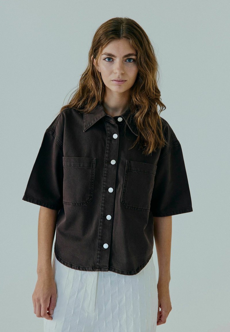 Chemise en denim noire à manches courtes avec un col, deux poches avant et des boutons blancs. Portée sur une jupe blanche texturée.