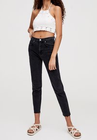 Jean noir taille haute, coupe slim, associé à un haut halter en crochet blanc. Le mannequin porte des sandales à lanières de couleur claire, en position debout.