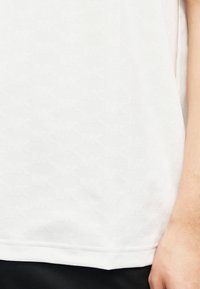 Gros plan d'un tissu blanc avec un motif en relief "MK" discret et répétitif, à côté du bras droit d'une personne et d'un pantalon noir.