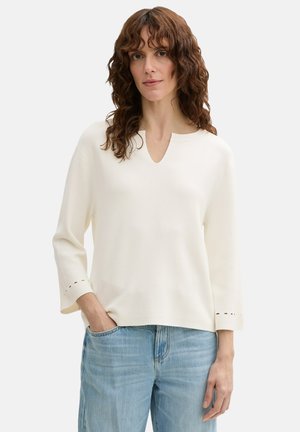 Pull crème à col en V, à manches trois-quarts, avec de petits détails découpés aux poignets ; associé à un jean bleu clair.