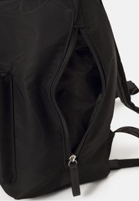 Zaino nero con esterno in nylon liscio e testurizzato, dotato di una tasca con zip e spallacci regolabili. L'interno è foderato in tessuto scuro.