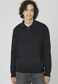 Pull-over en maille noir avec une demi-fermeture éclair, des poignets et une taille côtelés. Il présente un col et un tissu à texture douce. Porté par-dessus un haut léger.