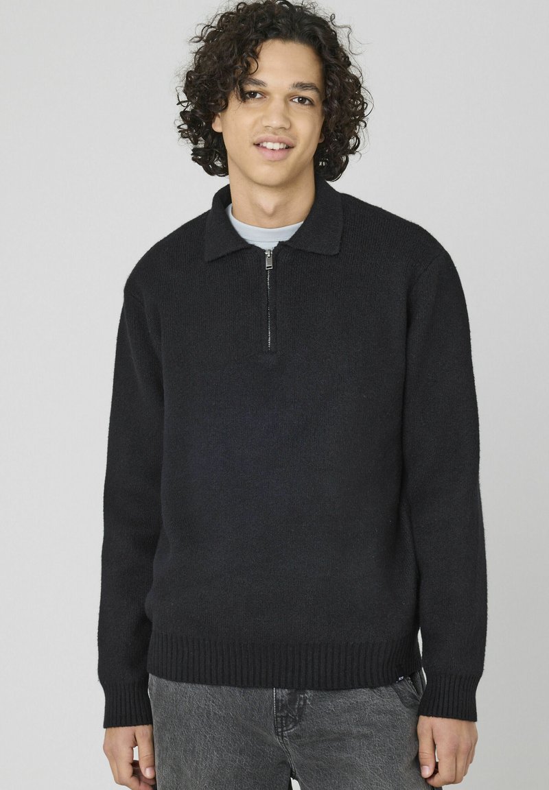 Pull-over en maille noir avec une demi-fermeture éclair, des poignets et une taille côtelés. Il présente un col et un tissu à texture douce. Porté par-dessus un haut léger.