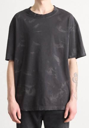 Person iført en løs sort tie-dye t-shirt og sorte bukser, med en slange-tatovering synlig på venstre underarm.