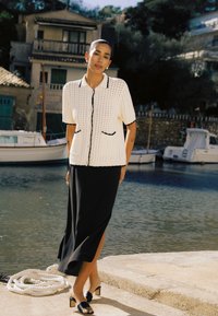 Femme debout près d'une marina, portant un cardigan en maille blanc à manches courtes et une longue jupe noire fendue sur le côté, avec des talons ouverts noirs.