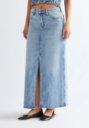 Kobieta ubrana w jasnoniebieską długą spódnicę jeansową z rozporkiem z przodu, w połączeniu z czarnymi balerinami i krótkim, paskowanym topem.